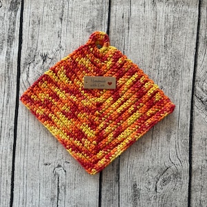 Könnte beinhalten: Ein gehäkelter Topflappen in Rot-, Orange- und Gelbtönen mit einem braunen Etikett, auf dem "Handmade with Love" steht.