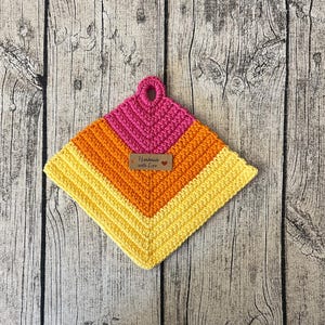 Peut inclure: Un dessous de plat crocheté en jaune, orange et rose avec une étiquette brune indiquant "Fait main avec amour".