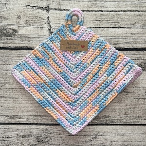 Könnte beinhalten: Ein gehäkelter Topflappen in Blautönen, Rosa und Pfirsich mit einem braunen Etikett, auf dem "Handmade with Love" und ein rotes Herz stehen.