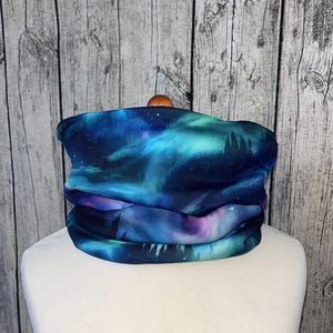 Può includere: Un scaldacollo con una vivace stampa galassia nei toni del blu, verde acqua e viola. Il tessuto presenta un design a cielo stellato con sagome di alberi. Lo scaldacollo è piegato e mostrato su una testa di manichino bianca.