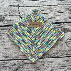 Könnte beinhalten: Ein gehäkelter Topflappen in helllila, gelb und blau. Der Topflappen hat einen Schlaufe zum Aufhängen und ein braunes Etikett mit der Aufschrift "Handmade with Love" und einem roten Herz.