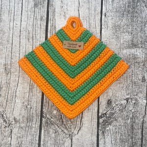 May include: Handmade crochet pot holder in a diamond shape. The pot holder features alternating stripes of orange and green yarn. A small tag with the words "Handmade with Love" is attached. The pot holder has a loop for hanging.