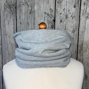 Op de afbeelding: Een lichtgrijze nekwarmer van zachte stof. De nekwarmer wordt getoond op een witte mannequin, met een houten bal bovenop. De achtergrond is een houten muur.