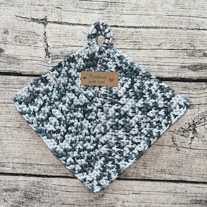Könnte beinhalten: Grauer und weißer gehäkelter Topflappen mit einer Schlaufe. Der Topflappen hat ein braunes Etikett mit der Aufschrift "Handmade with Love" und einem roten Herzen.