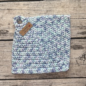 Op de afbeelding: Een vierkante, handgemaakte gehaakte pannenlap in tinten blauw, paars en wit. Het heeft een lus om op te hangen en een klein label met de tekst "Handmade with Love".