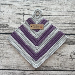 Op de afbeelding: Een grijze en paarse gehaakte ovenwant met een lusvormige handgreep. De ovenwant heeft een bruin label met de tekst "Handmade with Love" en een rood hart.