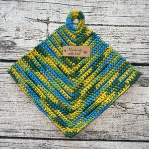 Op de afbeelding: Een groene, blauwe en gele gehaakte ovenwant met een lusvormige handgreep. De ovenwant heeft een bruin label met de tekst "Handmade with Love" en een rood hart.