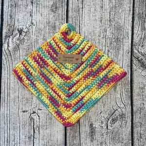 Könnte beinhalten: Ein gehäkelter Topflappen in Gelb, Rosa und Türkis mit einem braunen Etikett, auf dem "Handgefertigt mit Liebe" steht.