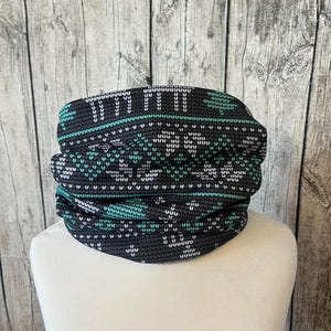 Puede incluir: Un calentador de cuello con estampado gris oscuro y verde azulado. El calentador de cuello presenta un patrón repetitivo de formas geométricas blancas y verde azulado. La tela está envuelta alrededor de una cabeza de maniquí blanca. El calentador de cuello está diseñado para usarse alrededor del cuello y la cara.