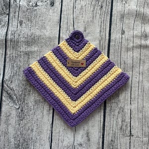Peut inclure: Un dessous de plat carré fait à la main au crochet, avec un motif en chevrons. Le dessous de plat présente des rayures alternées violettes et jaunes. Une petite étiquette indique "Handmade with Love" et comprend un symbole de cœur. Une boucle est en haut pour suspendre.