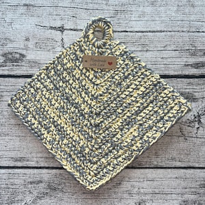 Op de afbeelding: Een grijze en gele gehaakte ovenwant met een lusvormige handgreep. De ovenwant heeft een bruin label met de tekst "Handmade with Love" en een rood hart.