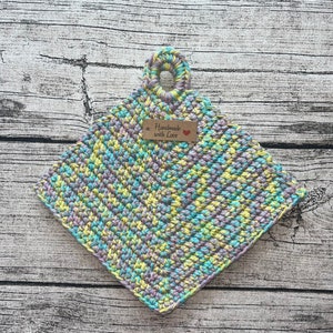 Op de afbeelding: Een gehaakte ovenwant in tinten van paars, groen en geel met een lusvormige handgreep. De ovenwant heeft een klein bruin label met de tekst "Handmade with Love" met een rood hart.