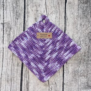 Peut inclure: Une manivelle de pot en crochet violet et blanc avec une étiquette brune qui dit "Handmade with Love".