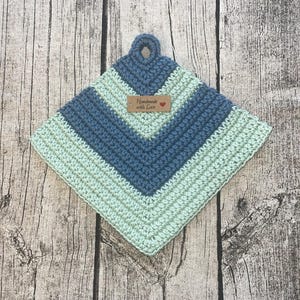 Op de afbeelding: Een handgemaakte, vierkante gehaakte pannenlap in tinten blauw en mintgroen. Het ontwerp heeft een chevronpatroon en een lus om op te hangen. Een klein label leest "Handmade with Love".
