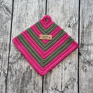Op de afbeelding: Een handgemaakte, vierkante gehaakte pannenlap in roze en olijfgroene tinten. De pannenlap heeft een lus om op te hangen en een klein label met de tekst "Handmade with Love". Het haakpatroon is een chevron ontwerp.