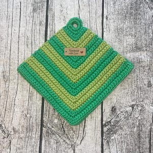 Op de afbeelding: Een handgemaakte, vierkante gehaakte pannenlap in groentinten. Het ontwerp heeft afwisselende strepen van licht- en donkergroen, met een lus om op te hangen. Een klein label leest "Handmade with Love".