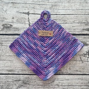Op de afbeelding: Een handgemaakte, vierkante gehaakte pannenlap in tinten paars, blauw en wit. De pannenlap heeft een zigzagpatroon en een lus om op te hangen. Een klein label leest "Handmade with Love."