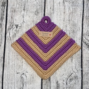 Op de afbeelding: Een gehaakte ovenwant met een bruin en paars gestreept patroon. De ovenwant heeft een lus aan de bovenkant en een klein label met de tekst "Handmade with Love".