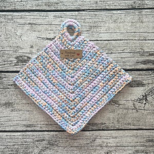 Op de afbeelding: Een vierkante, handgemaakte gehaakte pannenlap in pastelkleuren blauw, lavendel en perzik. Het heeft een lus om op te hangen en een label met de tekst "Handmade with Love". Ongeveer 20 cm vierkant.