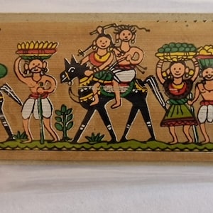 May include: A hand-painted wooden bookmark with a colorful scene of people riding animals. The bookmark features a brown background with black outlines and bright colors. The people are wearing traditional clothing and carrying baskets of fruit.
