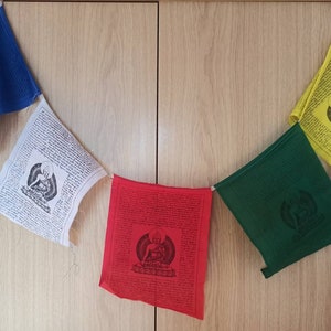 May include: A string of five prayer flags hanging on a wooden wall. The flags are blue, white, red, green, and yellow. Each flag features a different image of a Buddha and text in a foreign language.
