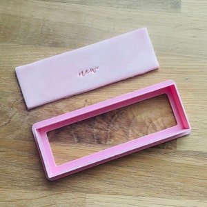 May include: A pink plastic cookie cutter in the shape of a rectangle with the word "new" embossed on the top. The cookie cutter is on a wooden surface.