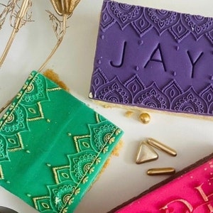 May include: Three decorated square cakes with intricate mandala designs. The cakes are purple, green, and pink. The purple cake has the name "JAY" written on it. The cakes are decorated with gold sprinkles.