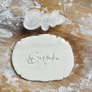 May include: A clear plastic cookie cutter and a piece of white dough on a wooden surface dusted with flour. The cookie cutter and the dough are imprinted with the word "Bespoke" in a cursive font.