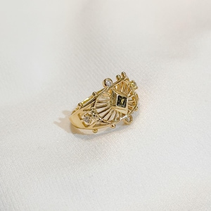 May include: A gold-colored ring with an intricate design. The ring features a central square-cut, dark green gemstone, surrounded by a sunburst pattern and small, clear accent stones. The ring is set against a white background.