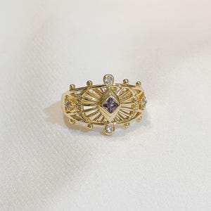 May include: Gold-toned ring with an intricate design. The ring features a central square-cut purple gemstone, surrounded by a sunburst pattern. Small, clear gemstones and decorative elements adorn the band, creating a detailed and elegant piece of jewelry.