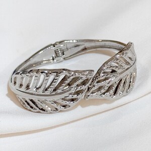 May include: A silver bangle bracelet with a leaf design. The bracelet is open and has a hinge clasp.