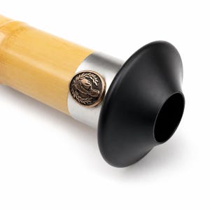 May include: A light-coloured bamboo flute featuring a silver-toned metal band near the end. The band is adorned with a circular bronze-coloured emblem depicting a hand within a laurel wreath. A smooth, matte black flared mouthpiece is attached.