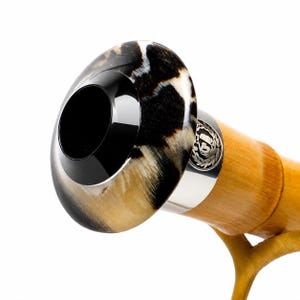 May include: A walking stick handle featuring a polished, rounded top with a black and white mottled pattern, a smooth black centre, and a silver-toned metal band embossed with a circular emblem, attached to a light brown wooden shaft.