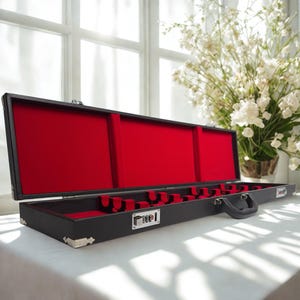 May include: A long, rectangular black carrying case with a hinged lid, featuring a vibrant red interior lining. The case includes a black handle, silver-toned metal corner protectors, and a combination lock mechanism on the front exterior.
