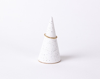 May include: A white ceramic ring holder shaped like a cone with black speckles. A gold ring is displayed on the holder.