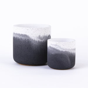 Può includere: Due vasi in cemento con effetto ombré, uno più grande dell'altro, con la parte superiore bianca e quella inferiore nera. I vasi hanno una texture punteggiata.
