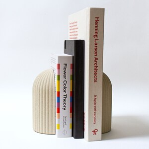 May include: A set of beige ceramic bookends with a ribbed texture, holding up three books. The book on the left is titled "Flower Color Theory" by  Droste and  Michael  Pfirsh. The book in the middle is black with a white spine. The book on the right is titled "Henning Larsen Architects" and has the text "It begins with curiosity" on the spine.