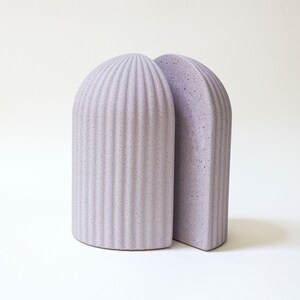May include: A pair of bookends in a light purple color. The bookends are made of a textured material and have a rounded, arched shape.