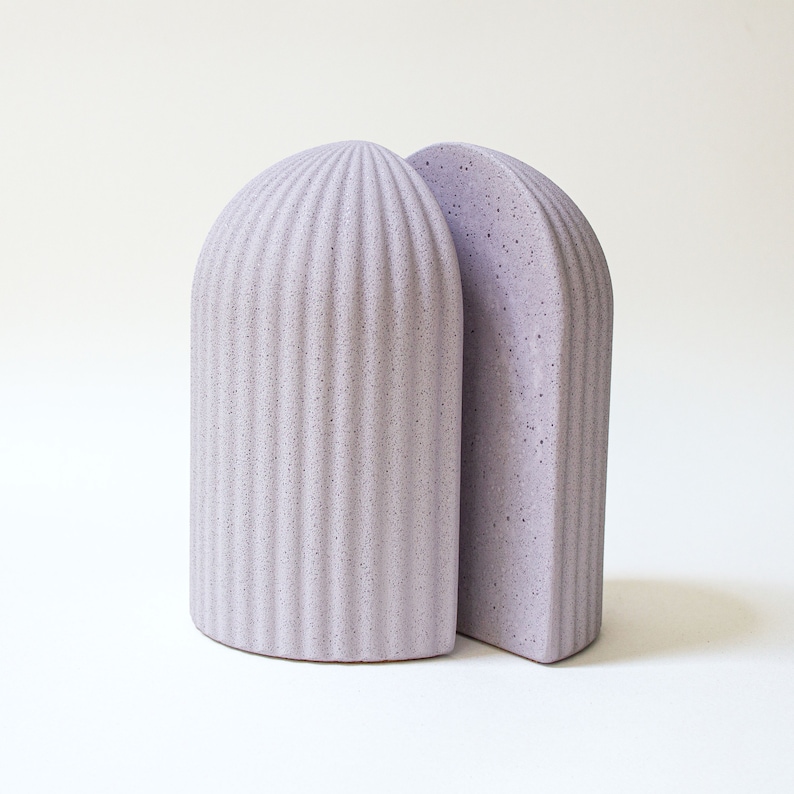May include: A pair of bookends in a light purple color. The bookends are made of a textured material and have a rounded, arched shape.
