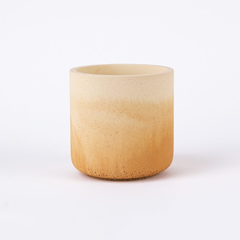 May include: A light brown ceramic planter with a white ombre effect. The planter has a textured surface and a round shape.