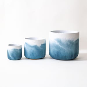 Puede incluir: Tres macetas de cerámica de diferentes tamaños. Cada maceta tiene una parte superior blanca y un diseño azul estilo acuarela que se desvanece hacia la parte inferior. Las macetas son cilíndricas y están sobre un fondo blanco.