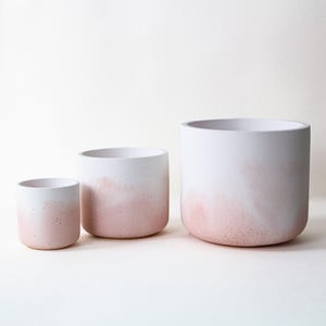 Puede incluir: Tres macetas cilíndricas de diferentes tamaños. Las macetas tienen una parte superior blanca que se desvanece en un tono rosa hacia la base. La maceta más pequeña está a la izquierda, seguida de una mediana y una grande a la derecha.