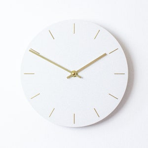 May include: A minimalist white wall clock with gold-colored hands and hour markers. The clock face is a solid white color with a textured surface. The hands are thin and elegant, and the hour markers are simple lines.