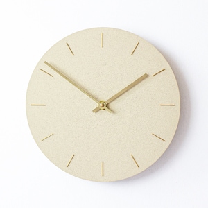 May include: A round, off-white clock with gold hands and gold hour markers. The clock face has a textured surface.