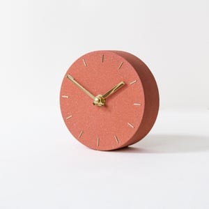 May include: A round, terracotta-colored desk clock with gold-colored hands and hour markers. The clock has a simple, minimalist design and is set against a white background. The clock's diameter is approximately 4 inches.