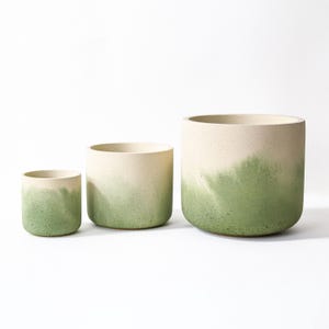 May include: Three cylindrical plant pots of different sizes. Each pot has a two-tone design, with a cream top that fades into a green hue at the bottom. The pots are arranged from smallest to largest, showing a gradient effect.