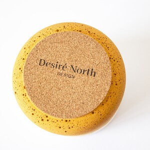 May include: The bottom of a yellow ceramic bowl with a cork base. The cork base has the text "Desire North Design" printed on it.