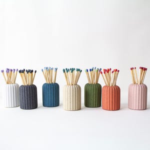May include: Seven decorative match holders in a row, each filled with matches. The holders are in a range of colours including white, navy, blue, beige, green, orange, and pink. The match heads are in various colours, adding to the visual appeal.