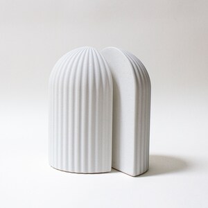 May include: A pair of white ceramic bookends with a ribbed design. The bookends are shaped like arches and are joined together at the top.