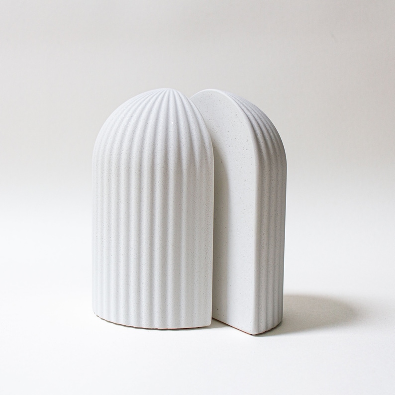 May include: A pair of white ceramic bookends with a ribbed design. The bookends are shaped like arches and are joined together at the top.